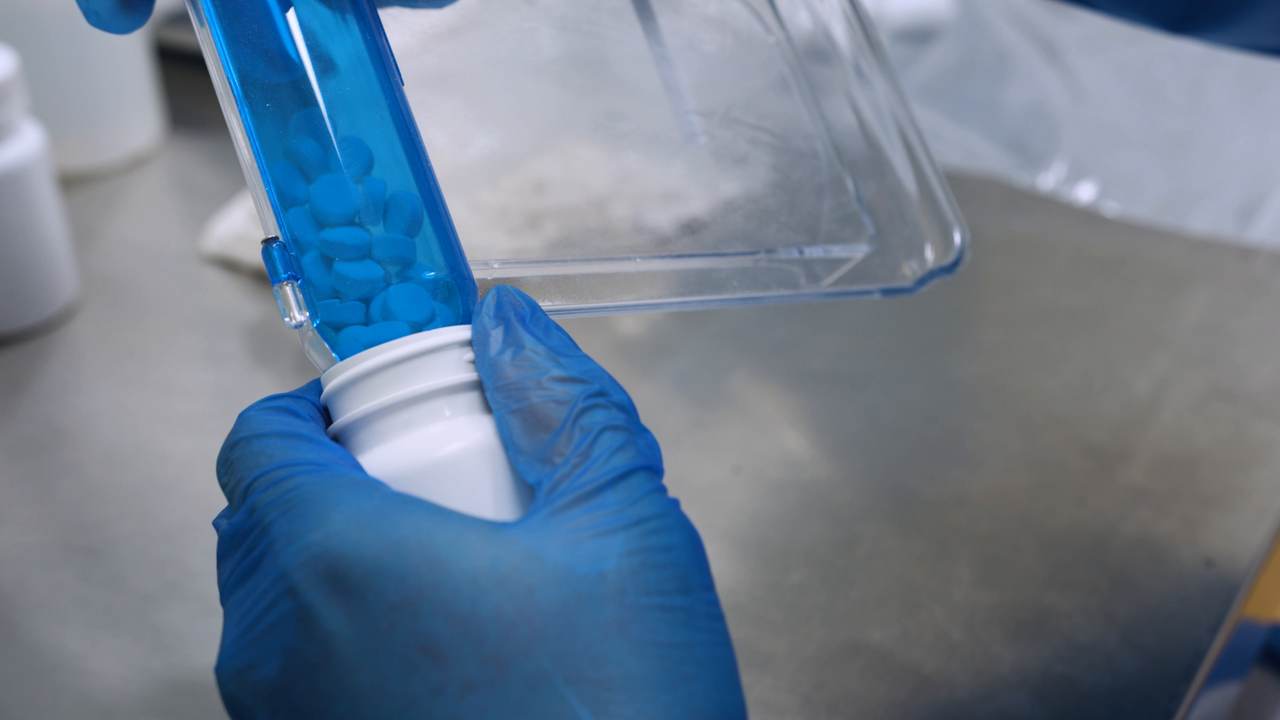 Gloved hands guide tablets from a blue tray into a plain white bottle